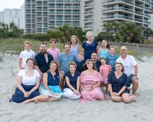 A group of people in coordinated navy and pink outfits pose together on a sandy beach with condominiums in the background.