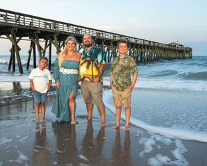 Family group stands together on beach at sunset with wooden pier and ocean waves in background.