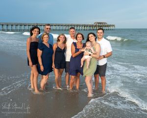 Group of people standing on beach with waves and pier in background at sunset.