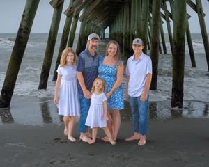 A family in blue and white outfits poses under a wooden pier at the beach on a cloudy day.