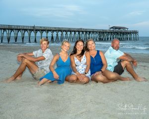 A group in colorful swimwear sits together on a sandy beach with a wooden fishing pier stretching into the ocean behind them.