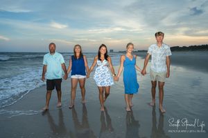A group of five people holding hands while standing on a beach at sunset with ocean waves in the background.