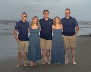 A group of people in coordinating blue and khaki outfits pose together on a cloudy beach at sunset.