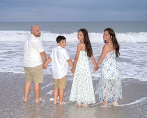 Family wearing coordinating white and blue outfits walking together along the ocean shoreline at sunset.