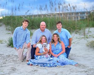 Family group wearing coordinated blue outfits poses together on sandy beach dunes at sunset.