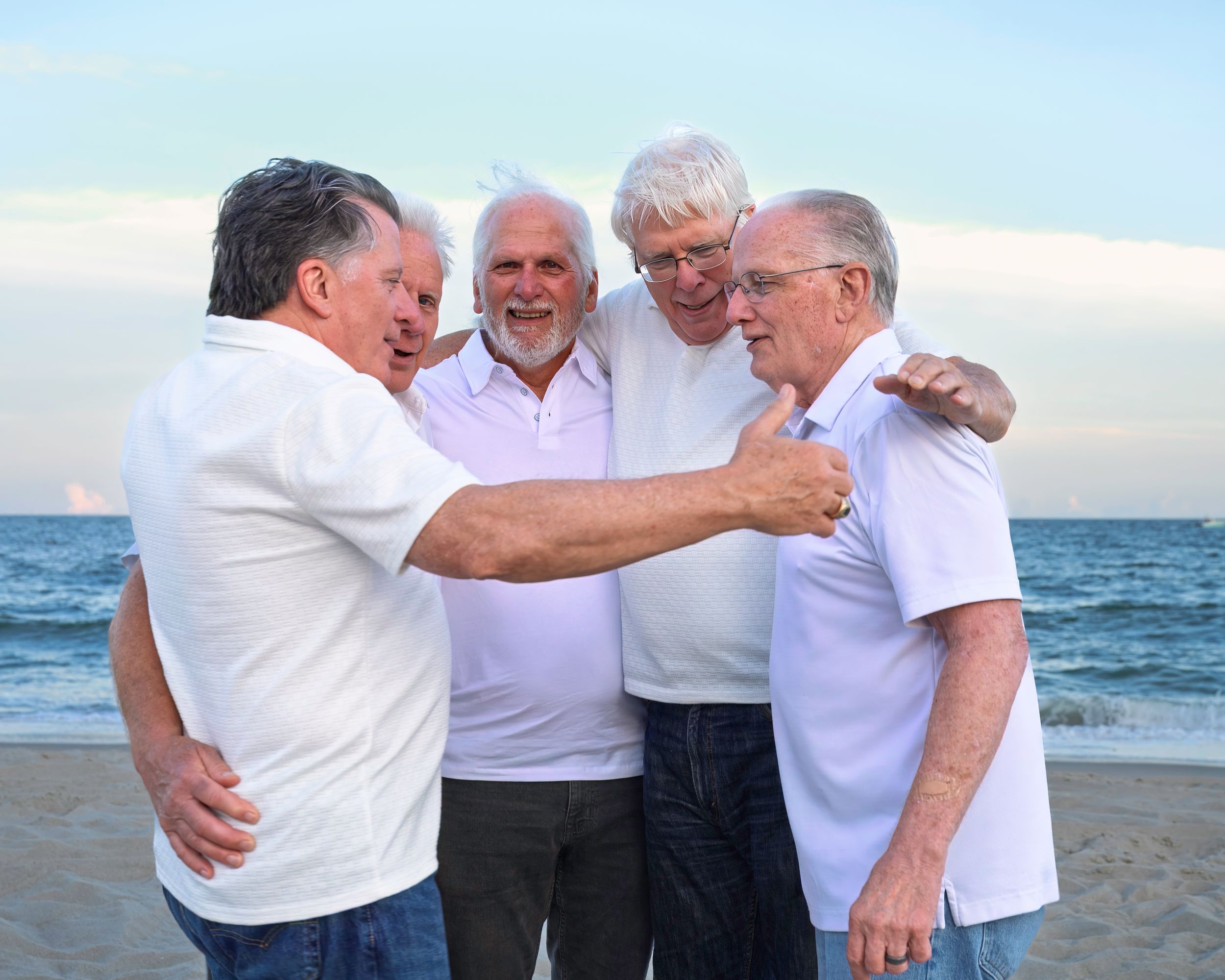 Group of senior friends in white shirts laughing and embracing while walking together on a beach at sunset.