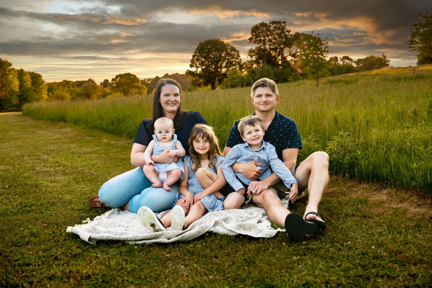A family relaxes together on grassy hill during golden hour sunset in matching light blue and white outfits.