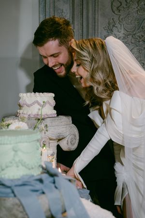A joyful wedding couple in white dress and dark suit laughing together over their tiered wedding cake.