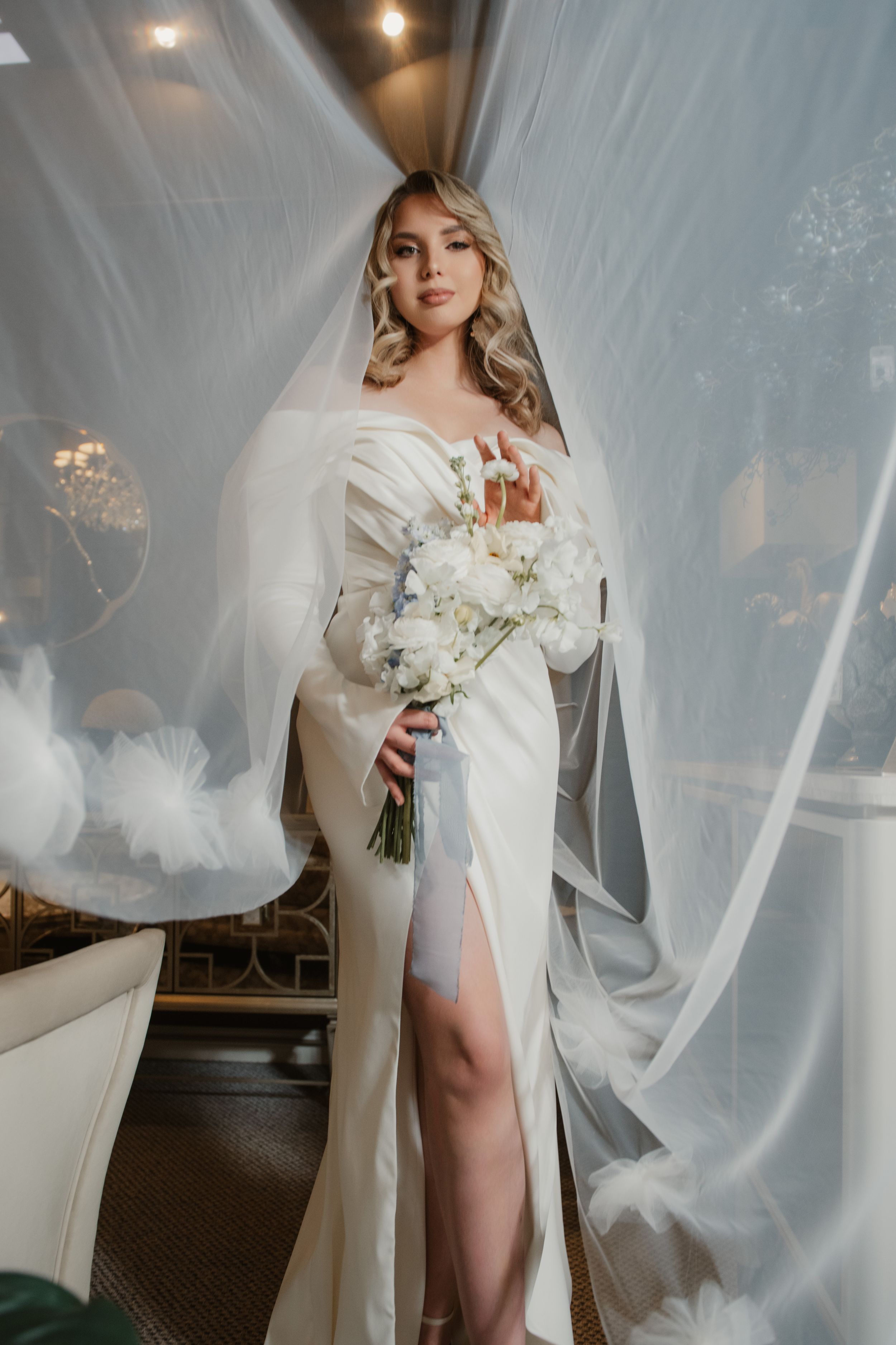 A bride in an elegant white dress poses gracefully with sheer flowing fabric draped around her in a dramatic indoor setting.