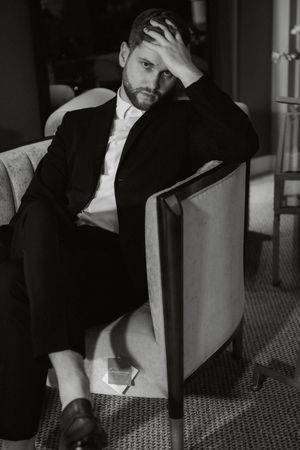 Man in black suit and white tie leans against industrial surface in dramatic black and white photo series.