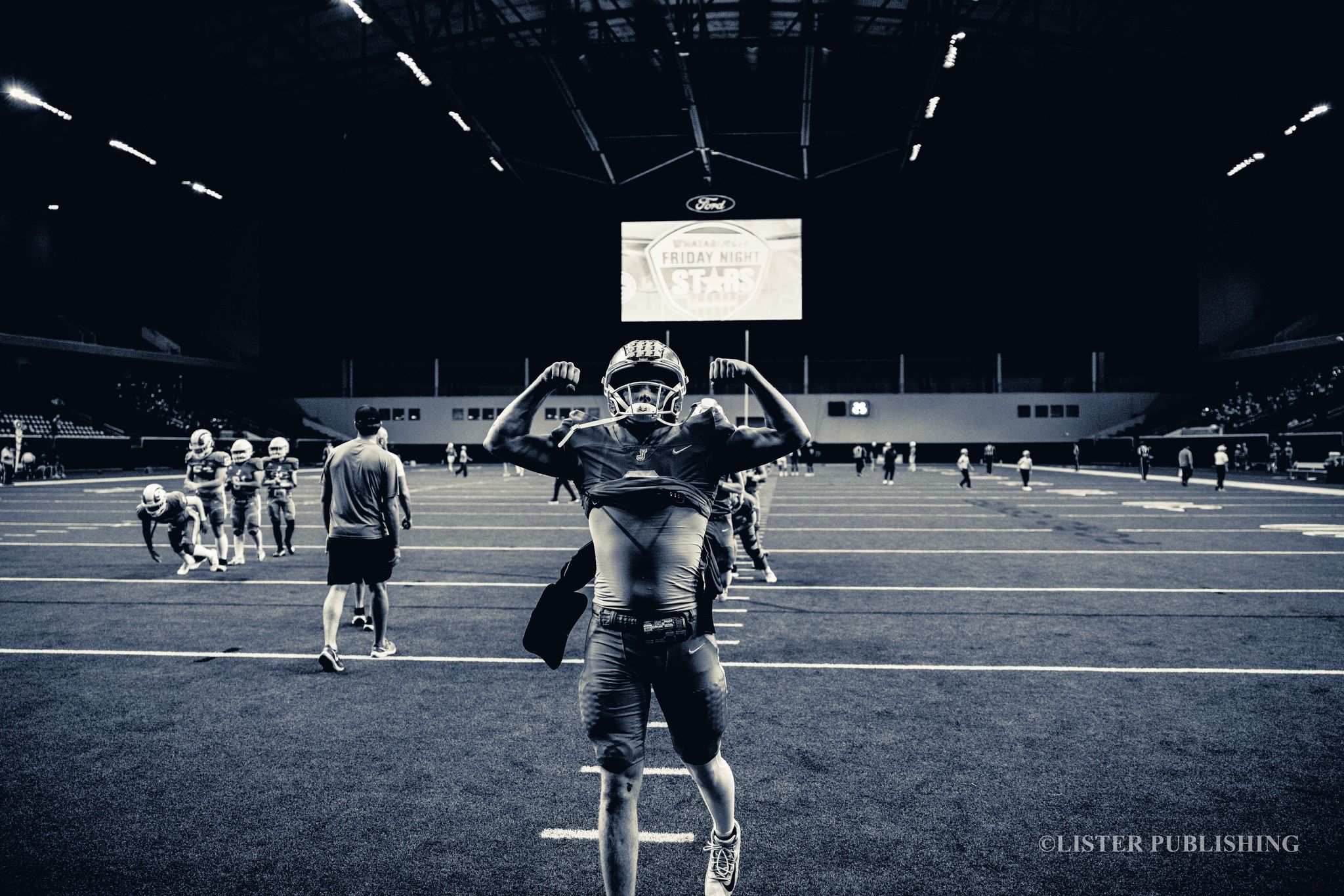 Black and white photo of a drum corps member performing on a football field at an indoor stadium.