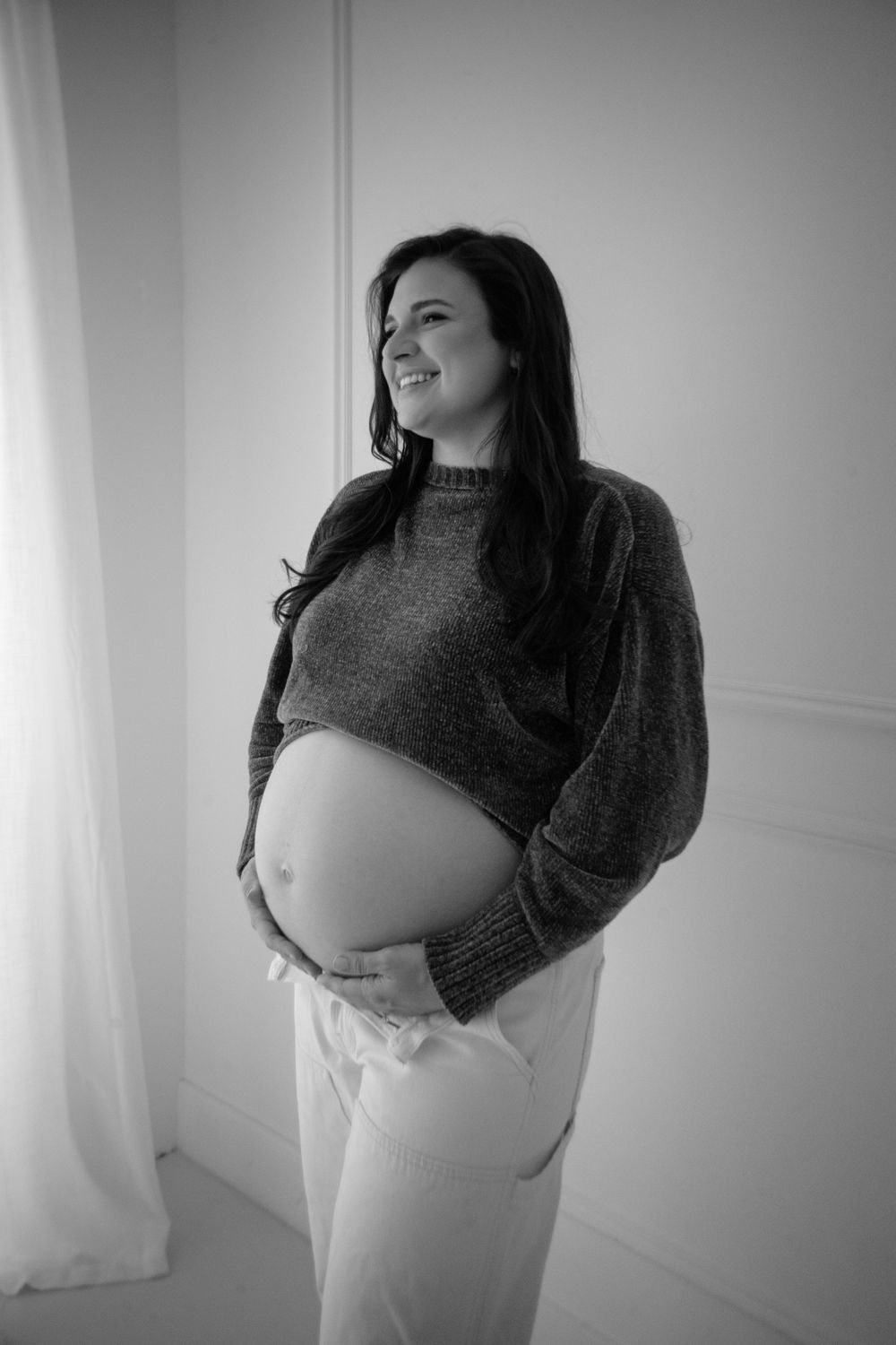 Black and white portrait of an expectant mother in a sweater and white pants cradling her baby bump.