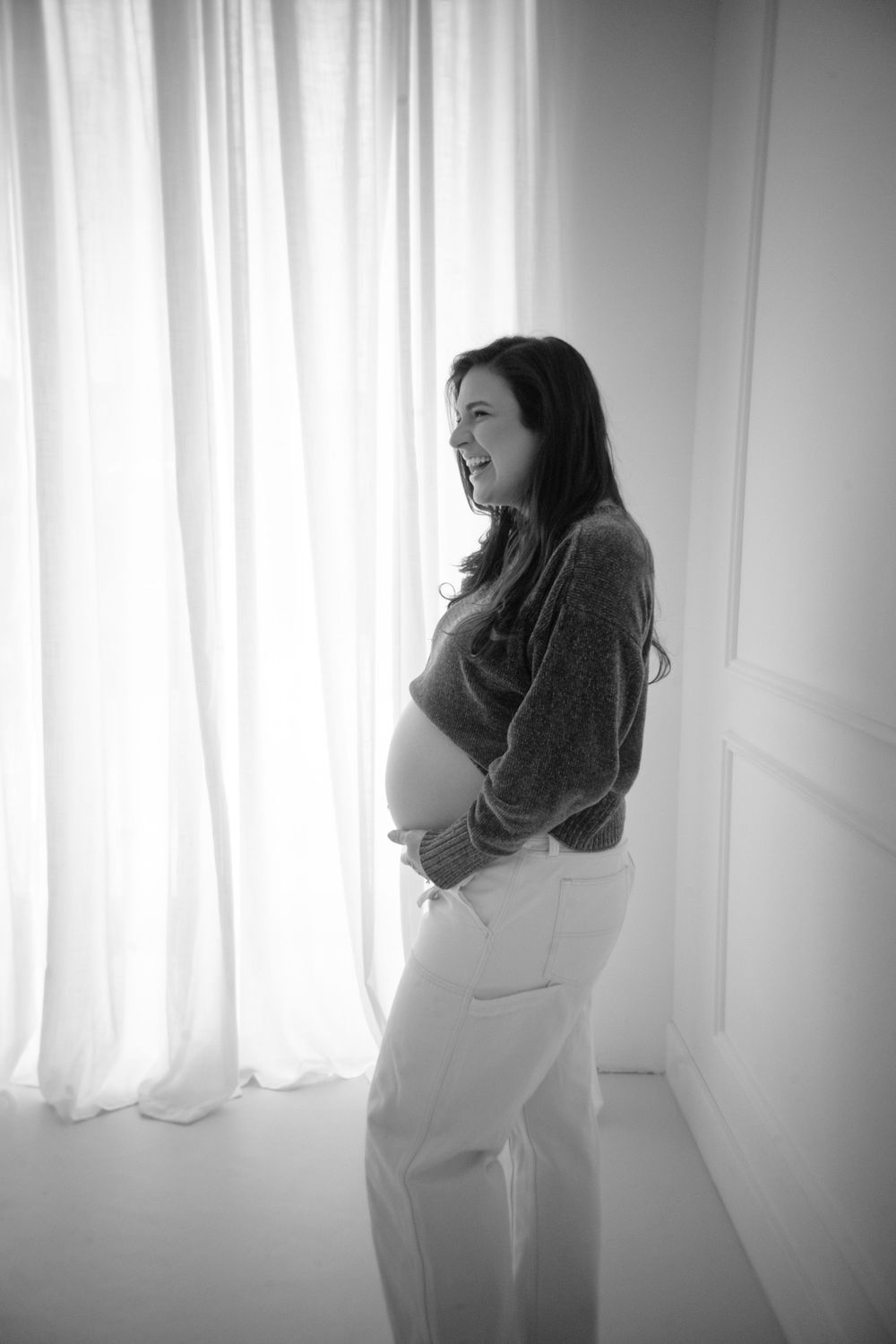 A joyful side-view maternity portrait in black and white by a window.