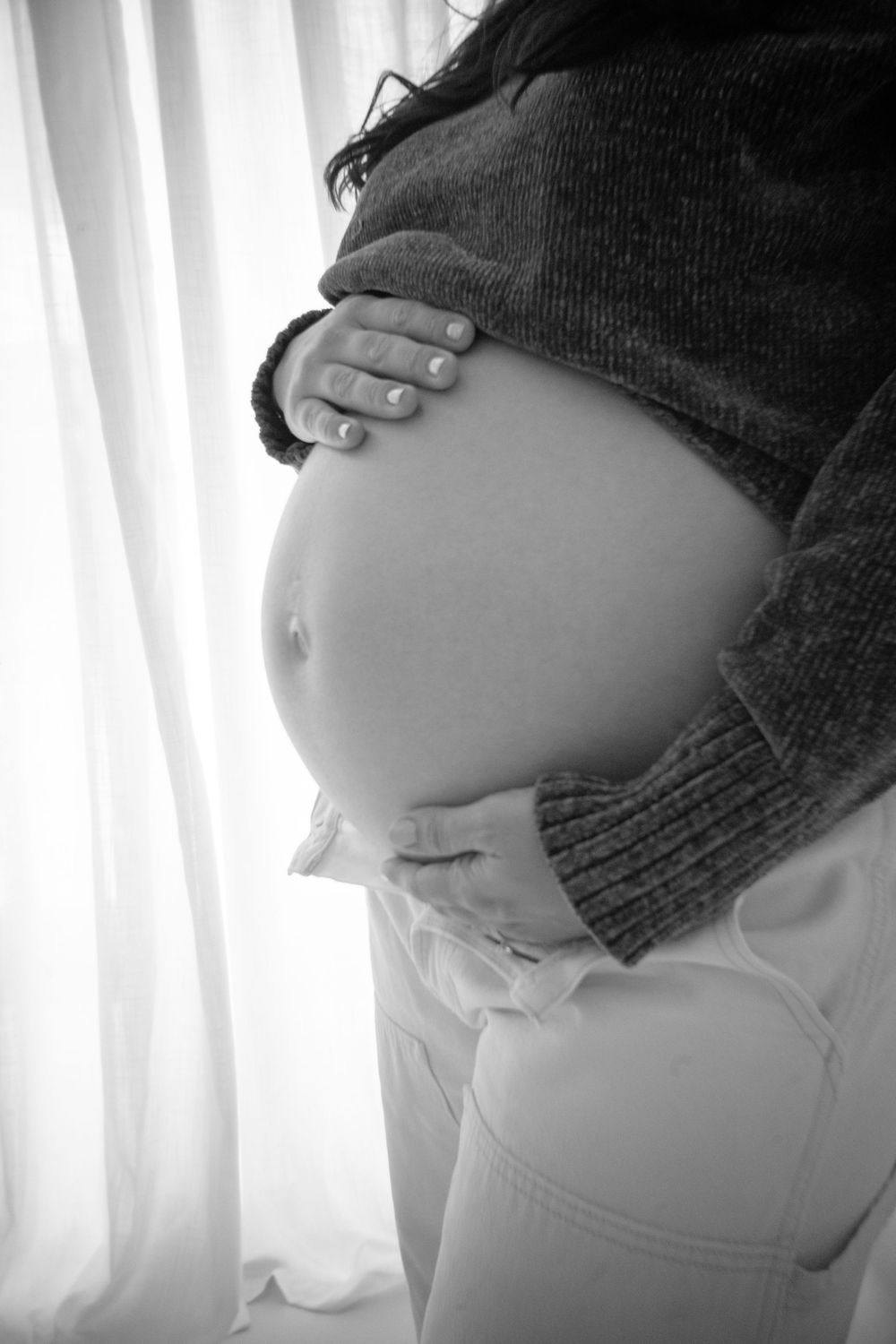 Black and white profile view of a pregnant woman's belly wearing a dark sweater against a light curtained background.
