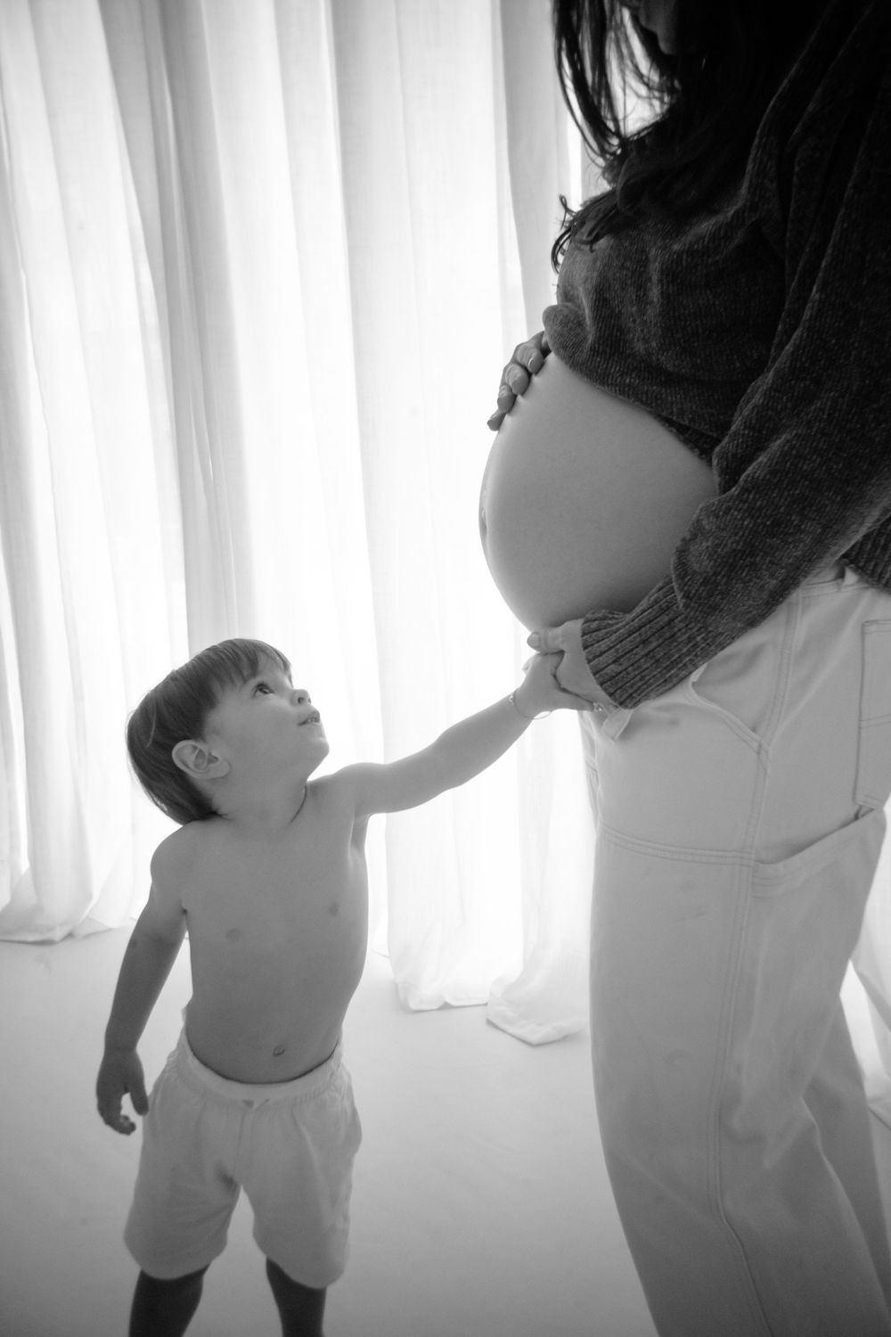 A intimate black and white photo of a toddler reaching up to touch a pregnant belly.