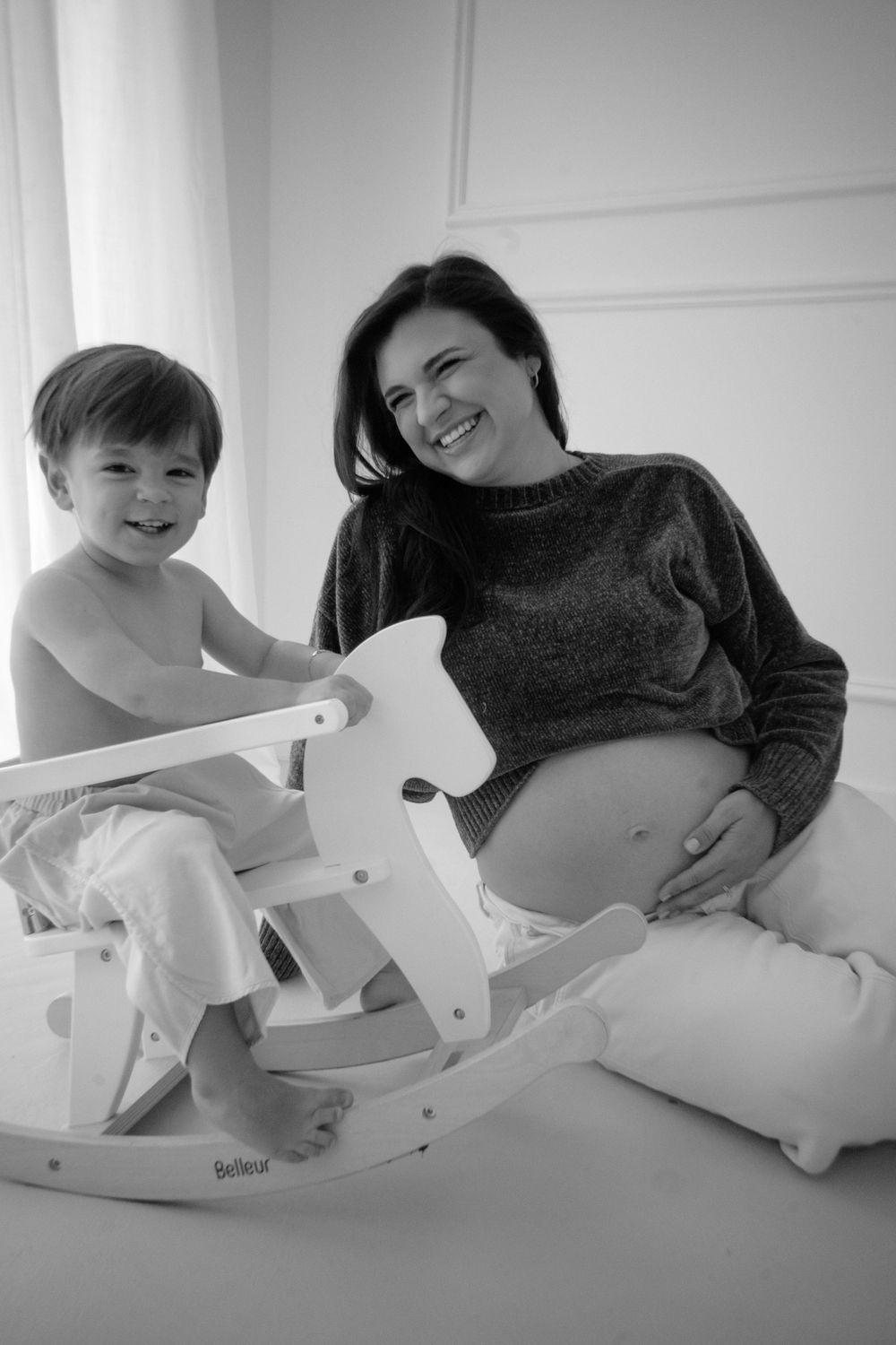 Heartwarming maternity photo showing child on rocking horse while expectant mother smiles from nearby.