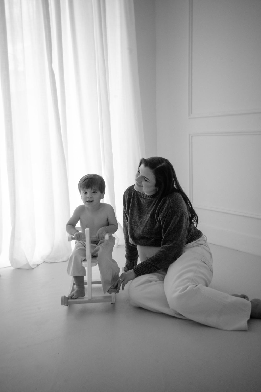 A toddler rides a rocking horse while an adult sits nearby in a minimalist white room setting.