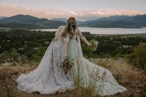 Wide shot of LGBTQ+ brides kissing in front of an alpine lake at sunset in Colorado.