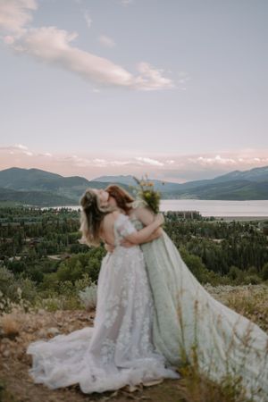 The two brides embrace, Emma kissing Victoria on the neck, in front of an alpine lake at sunset in Colorado.