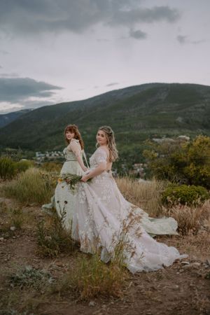 Emma leads her partner Victoria by the hand along a trail in Colorado, their dresses trailing behind.