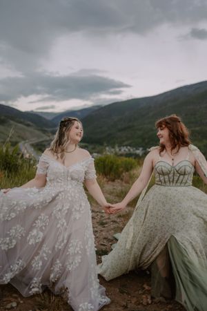 LGBTQ+ brides hold hands and swing their dresses on a trail overlooking Silverthorne, Colorado.