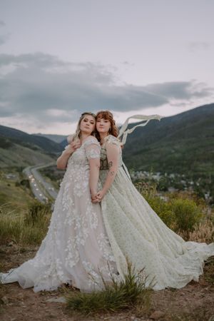 Brides Emma and Victoria pose, holding hands, on a mountain trail in Colorado.