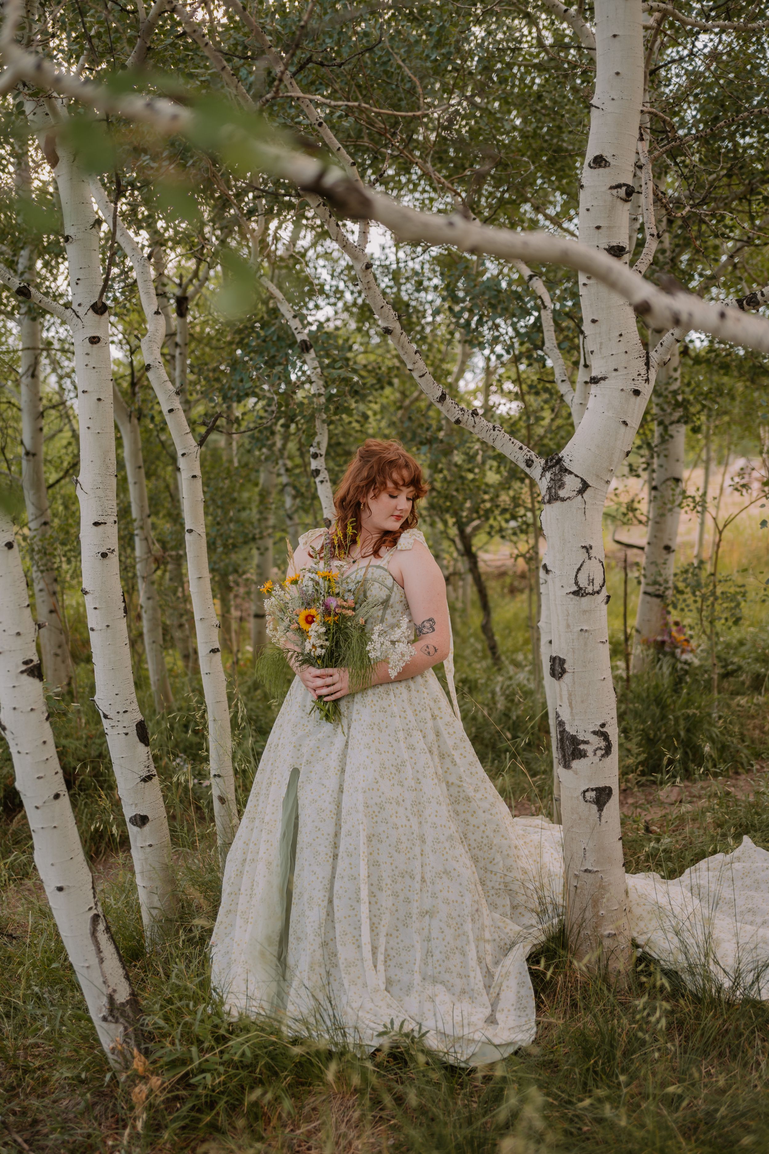 Emma, a bride with curly red hair, stands between aspen trees and looks down at her pale green wedding dress.