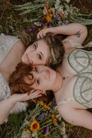 Shot from above of brides Victoria and Emma lying on the ground, heads next to each other, looking into the camera.