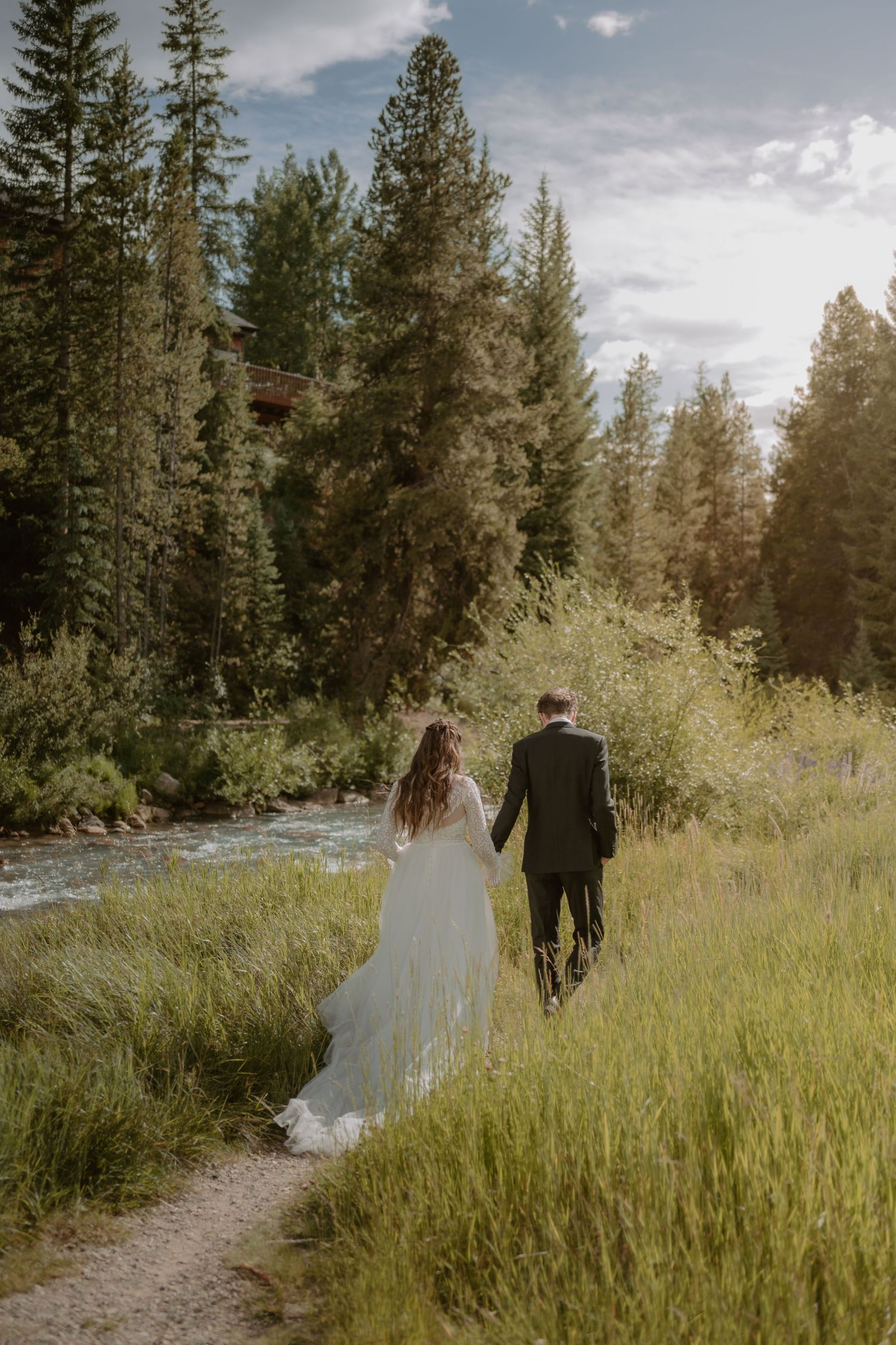 Newlyweds walking hand in hand along mountain stream path at sunset.