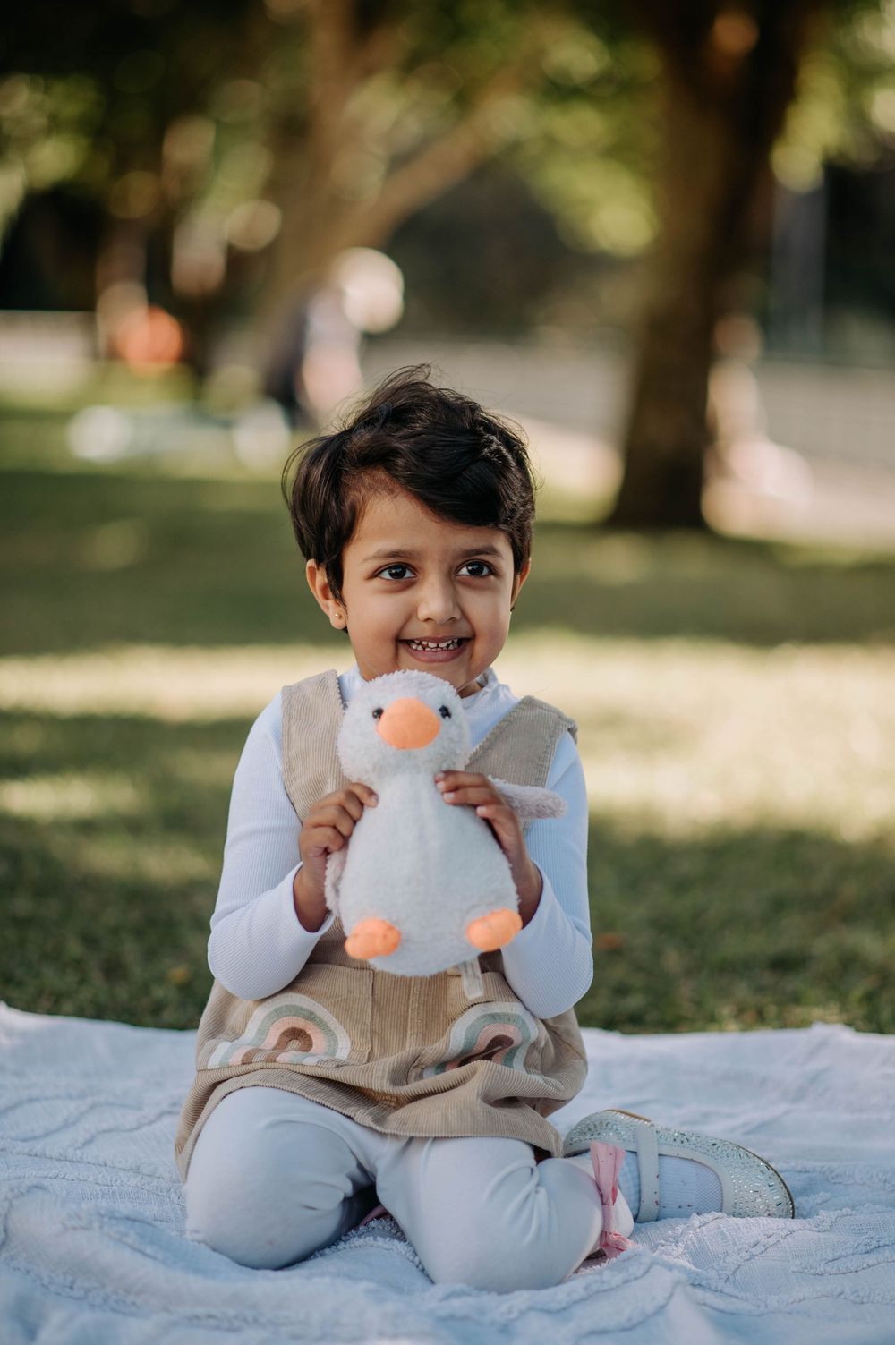 A young child sits on a blanket outdoors holding a plush penguin toy and smiling at the camera.