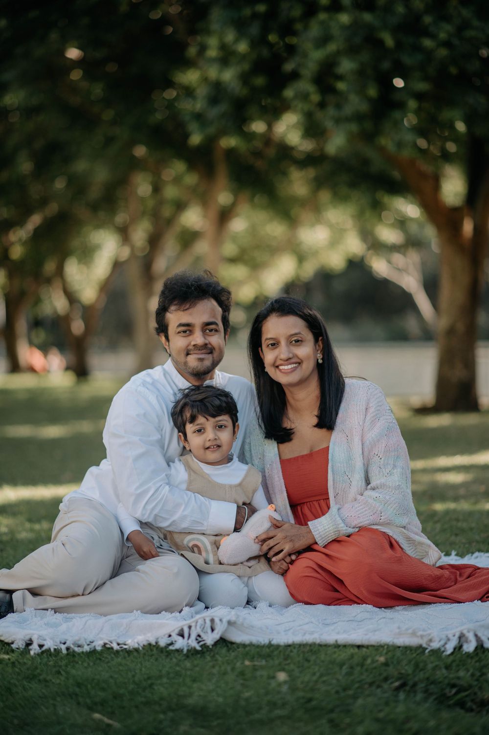 A family wearing white and red outfits lies on a picnic blanket on green grass under trees in a park setting.