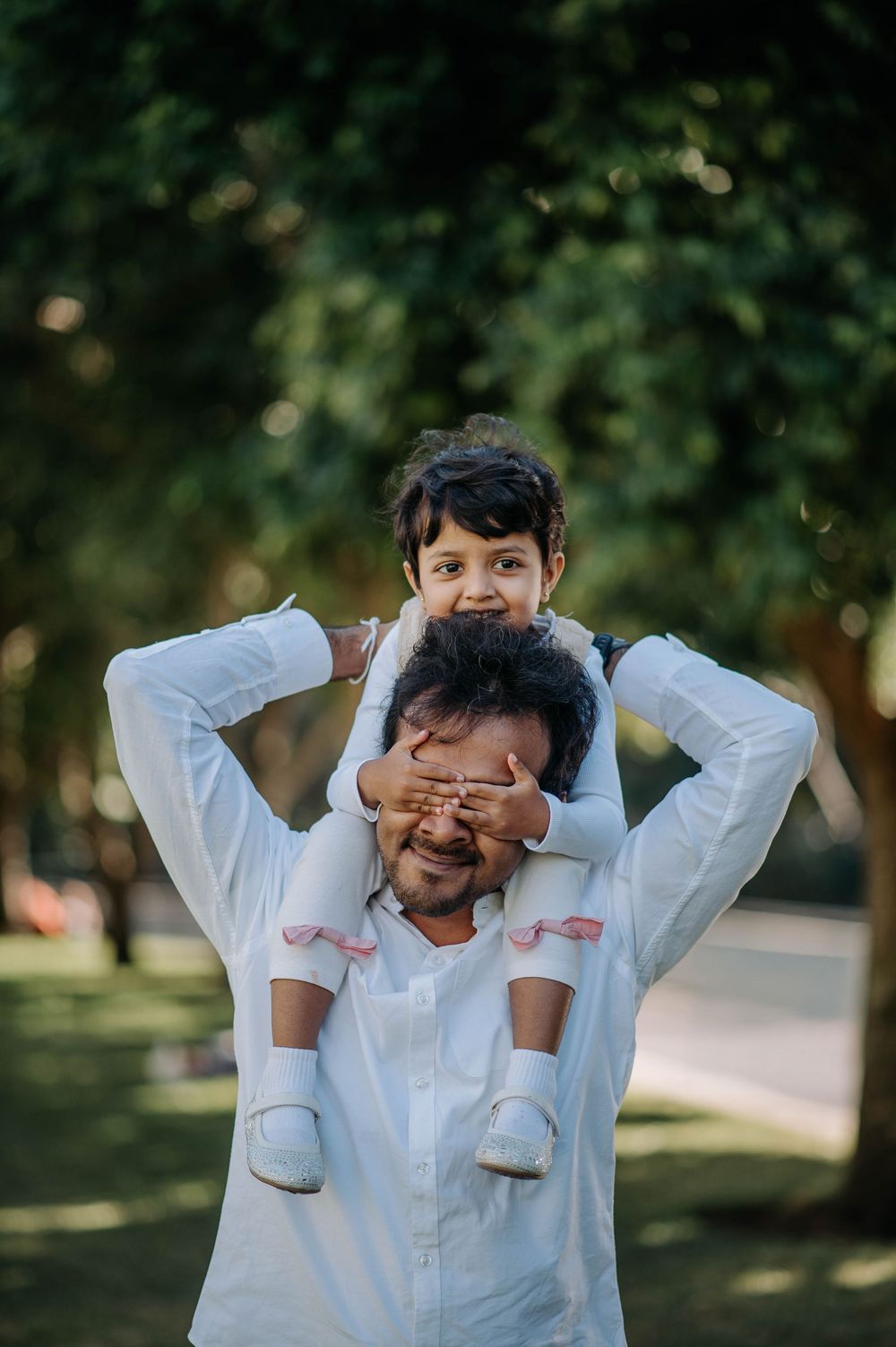 Playful outdoor family moment between parent and child in white outfits against blurred green park background.