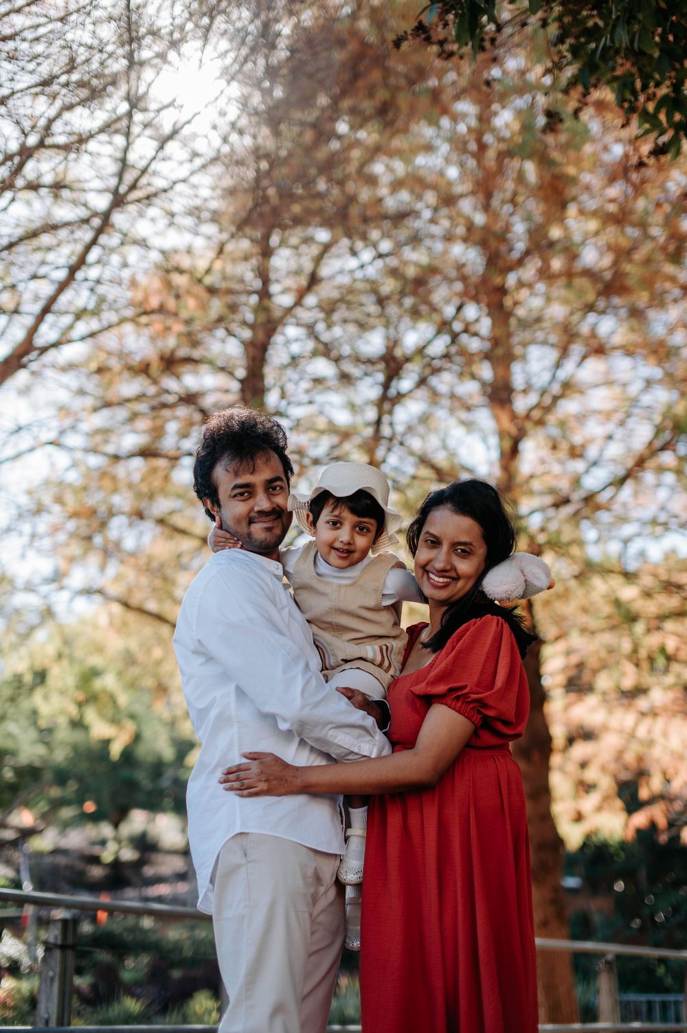 A family of three embraces outdoors during autumn with fall foliage in the background.