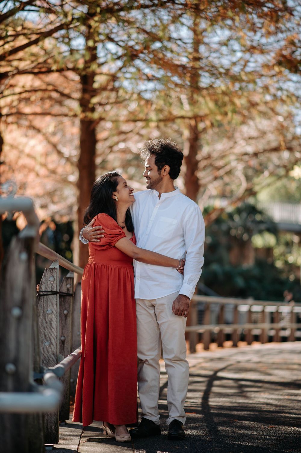 A couple embraces on a walkway with autumn trees in the background, wearing red and white outfits.
