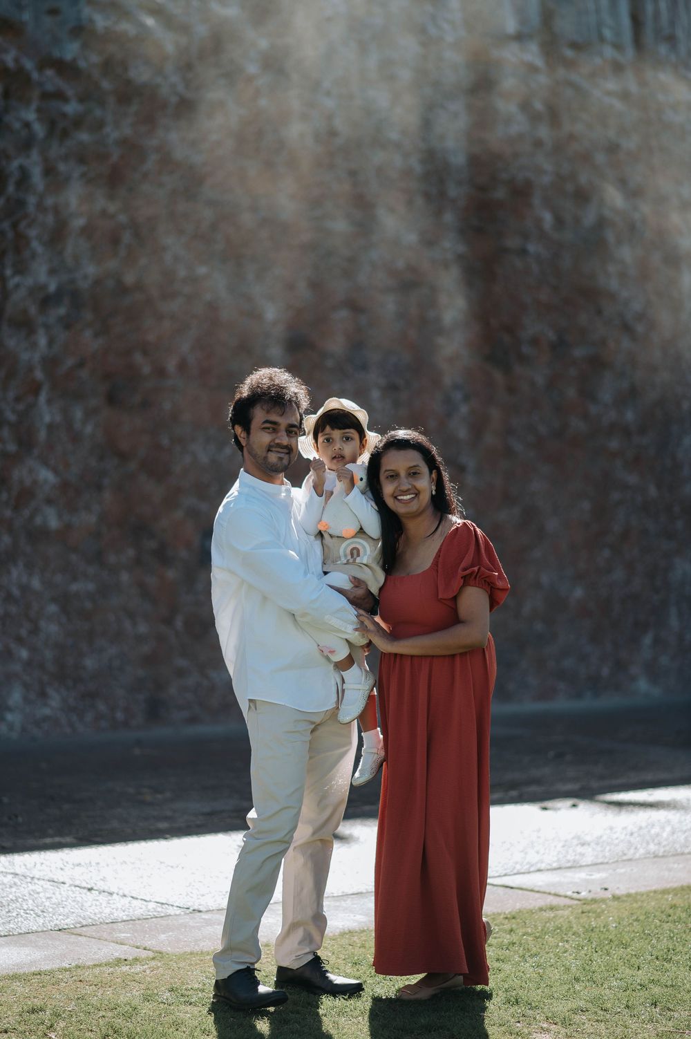 A family with a small child poses outdoors in front of a stone wall on a sunny day.