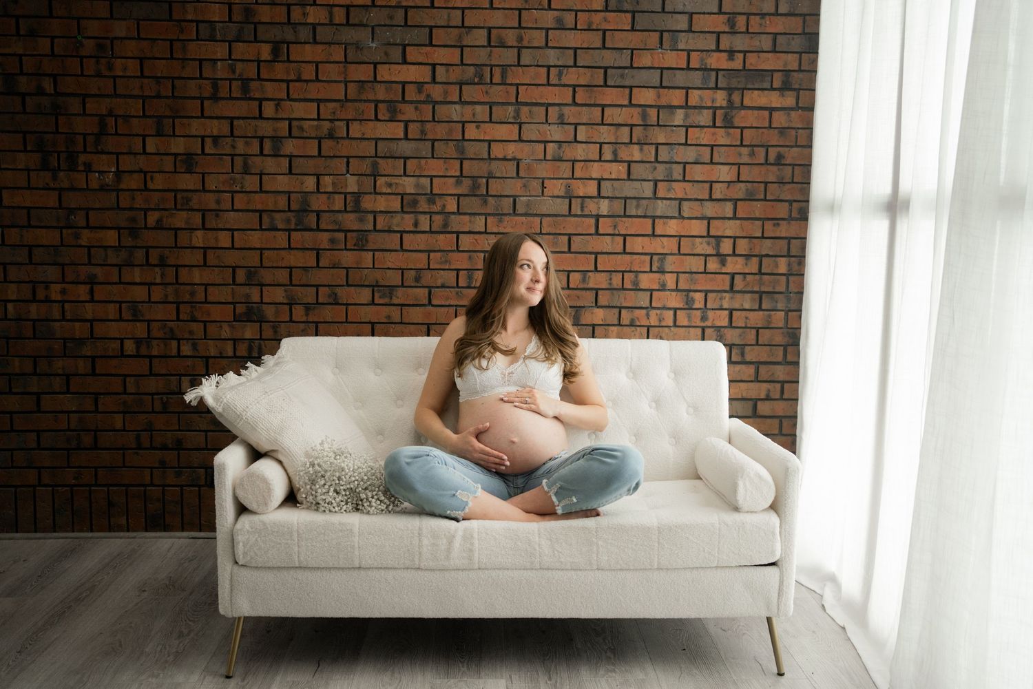 Person in casual attire rests on a beige sofa against a brick wall near a bright window.