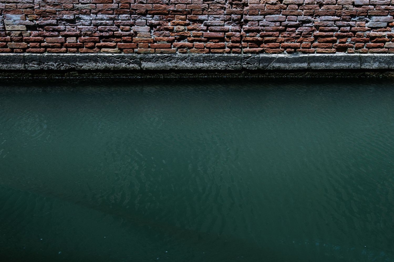 Dark green canal water meets an aged brick wall creating a minimalist urban composition.
