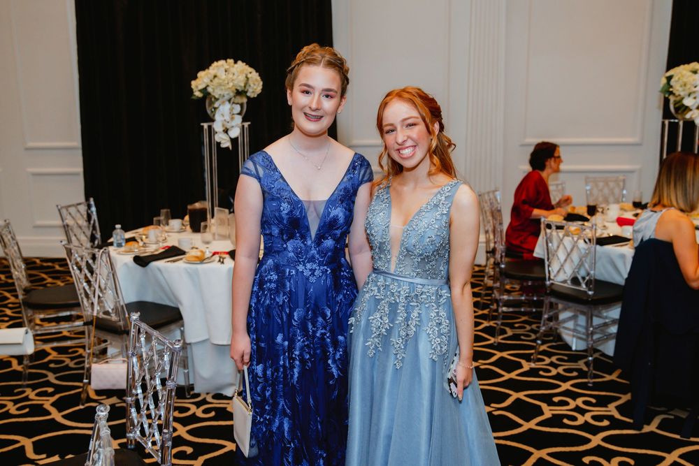 Two people wearing elegant evening gowns, one in royal blue and one in light blue, pose together at a formal event.