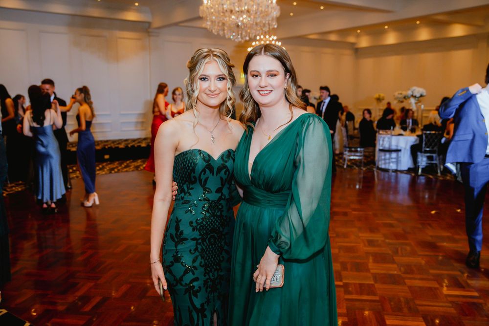 Two women pose together at a formal event wearing elegant emerald green evening gowns on a wooden dance floor.