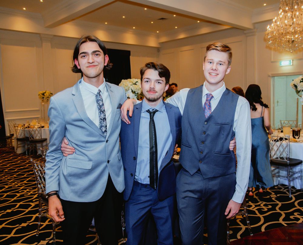 Groups of guests in formal attire pose and smile together during an elegant indoor event with chandeliers and carpeting.