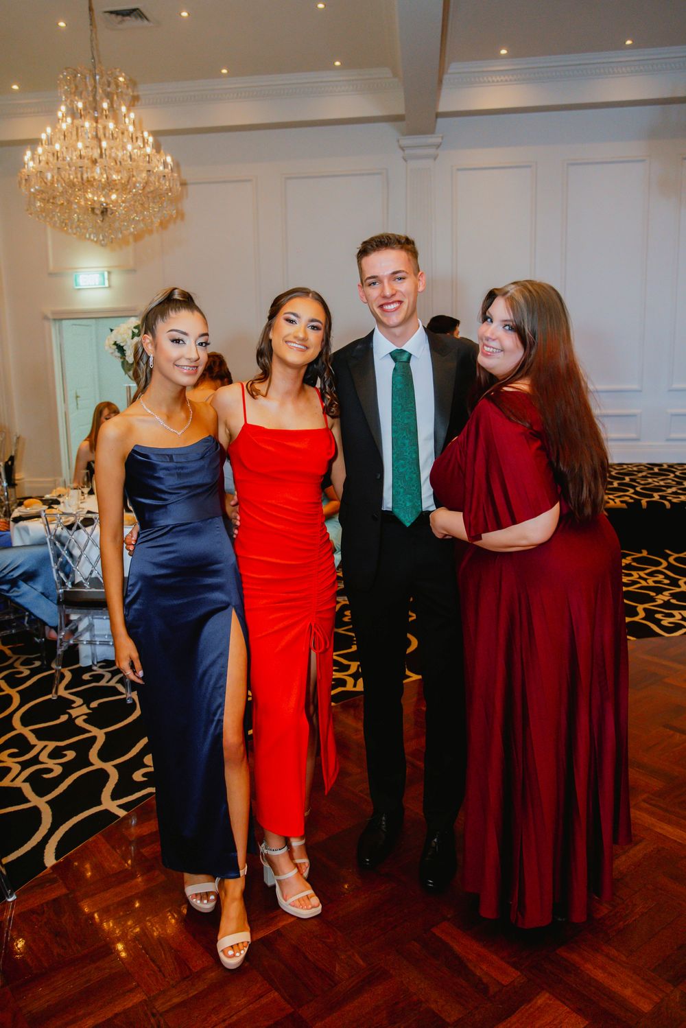A group in formal attire including navy, red and burgundy dresses pose together at an elegant reception venue with chandeliers.