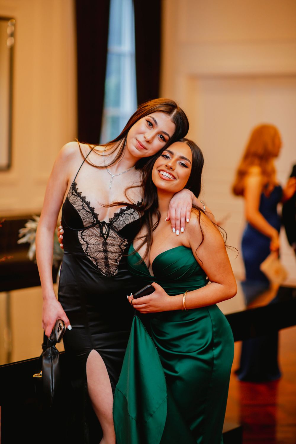 Two friends pose together at a formal event, one wearing an elegant emerald green gown and the other in a black lace dress.