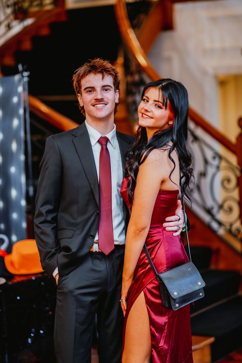 A couple in formal attire poses on a staircase, with the woman wearing a red satin dress and the man in a black suit.