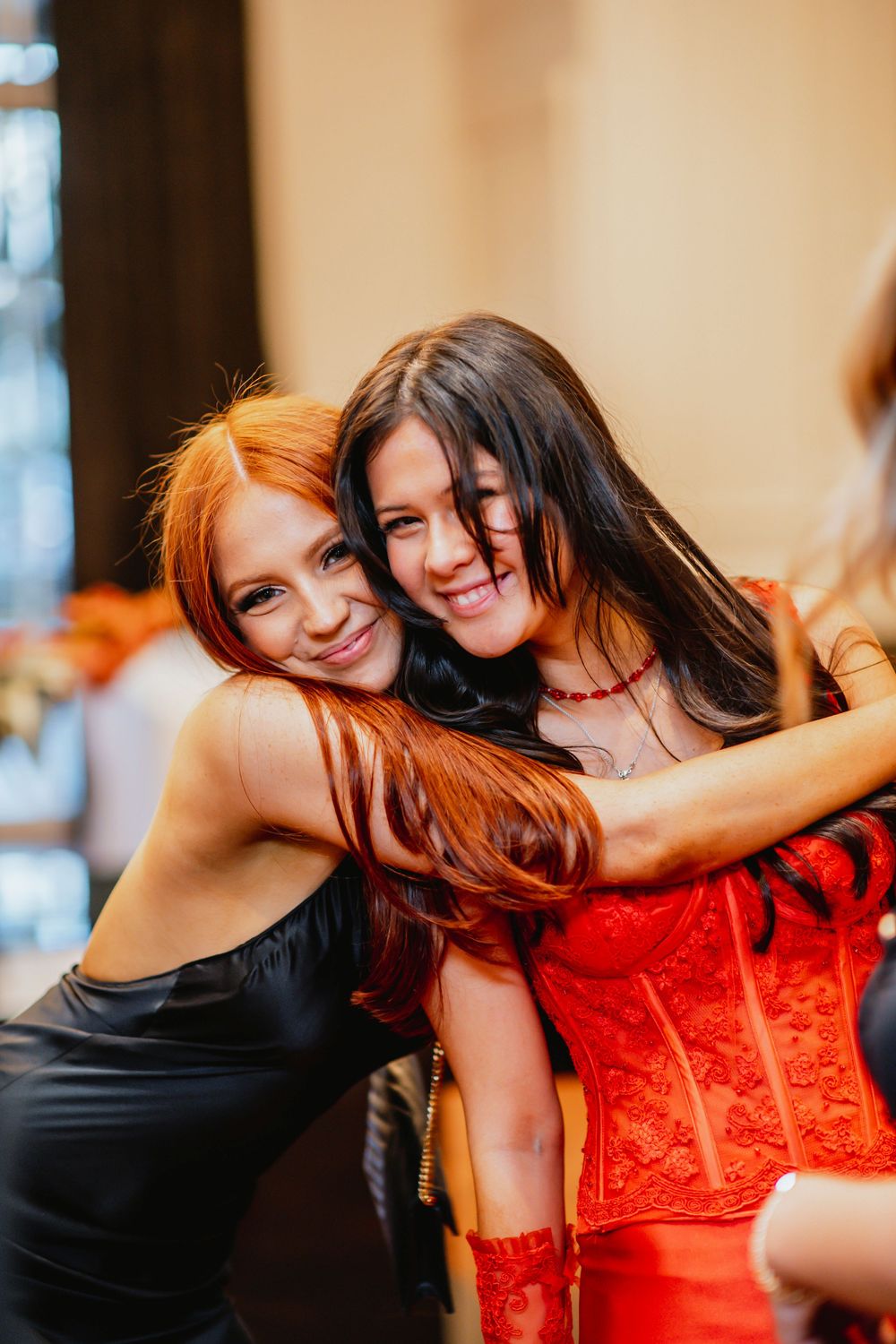 Two friends share a joyful embrace at a party, one wearing a black dress and another in a red corset.
