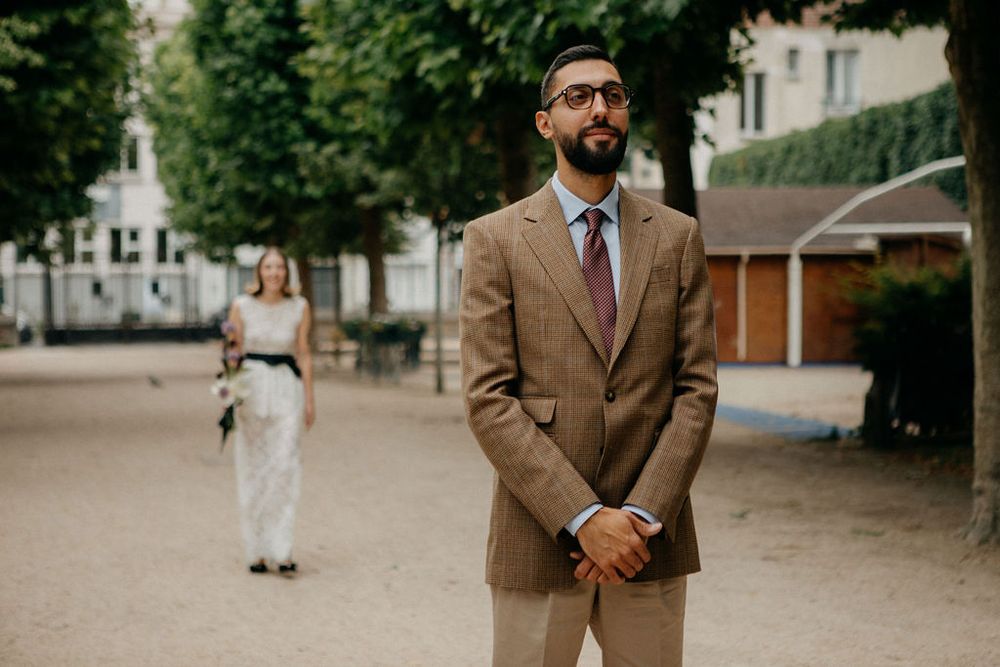 Groom in a tan suit standing in a tree-lined square during a first look wedding moment in Paris.
