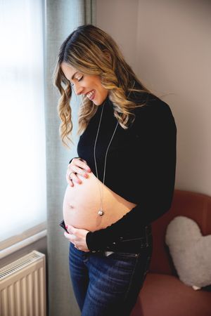 Expectant woman in black crop top and jeans smiles while touching her bare pregnant belly near a window. At a London maternity session.