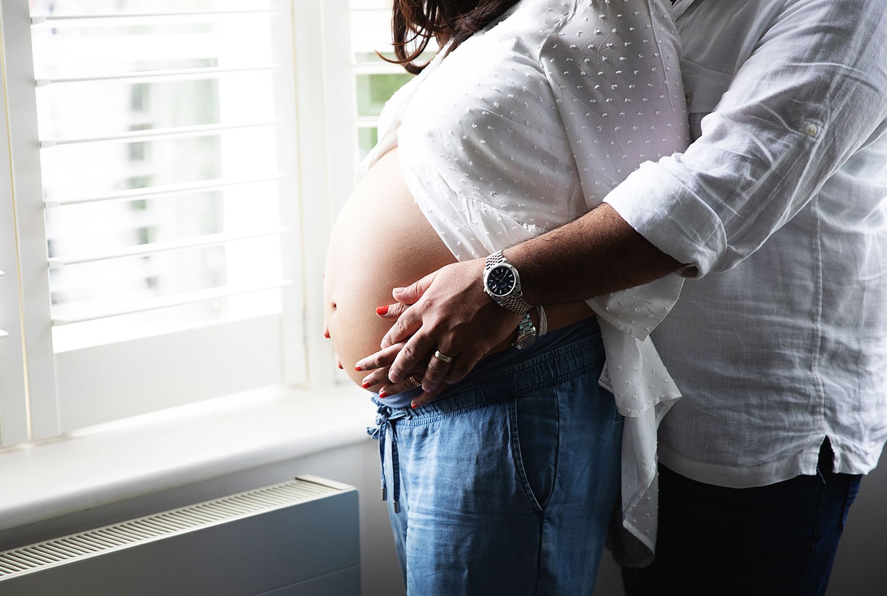 Person in a white button-down shirt and jeans poses casually by a bright window with natural lighting. During a London maternity session