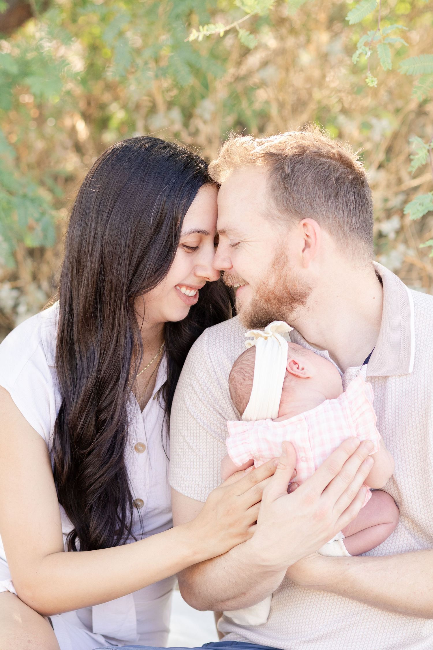 Dad holding newborn daughter smiling forehead to forehead with mom underneath a tree.