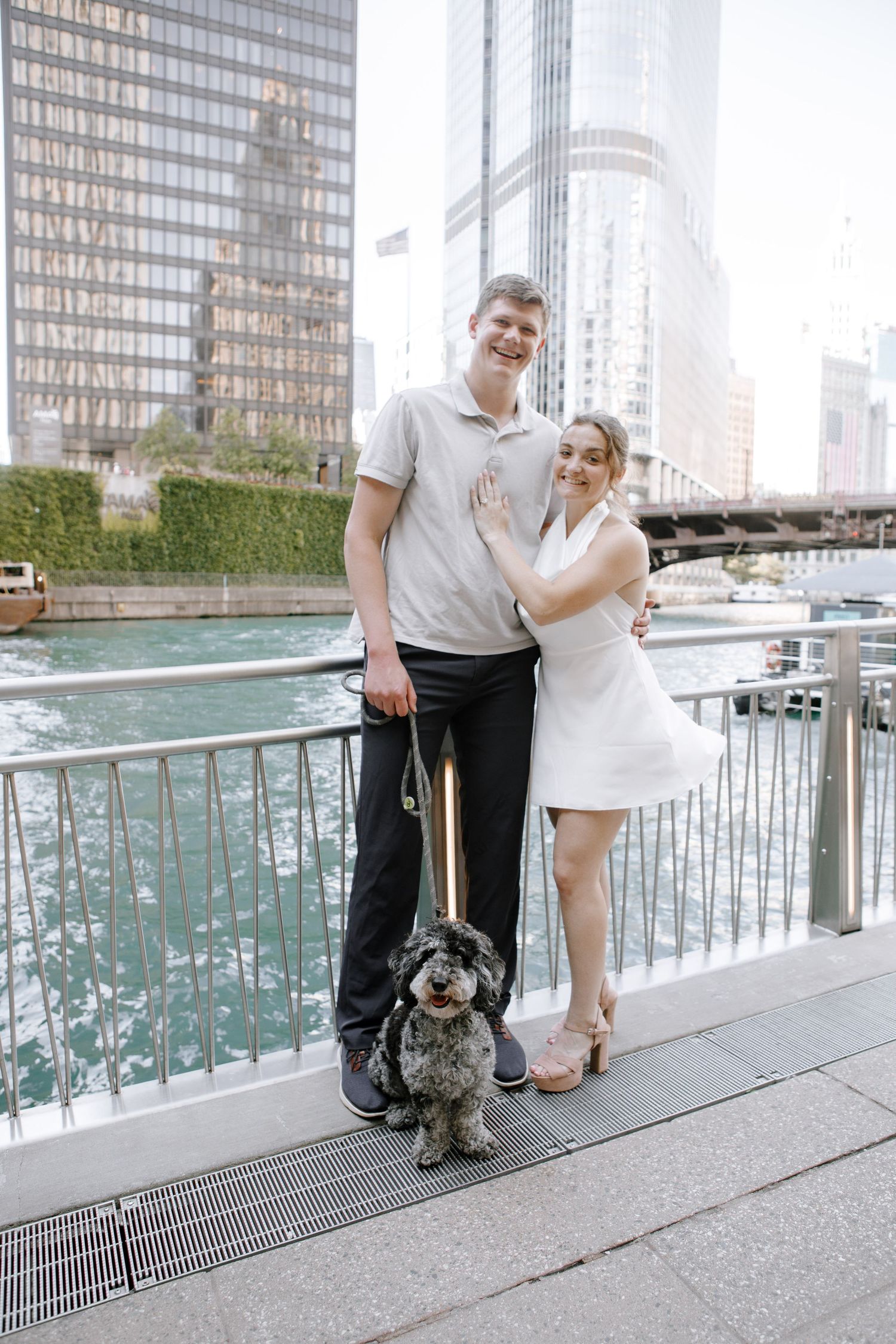 Couple poses with their small gray dog along a waterfront railing in downtown Chicago near skyscrapers and fountains.