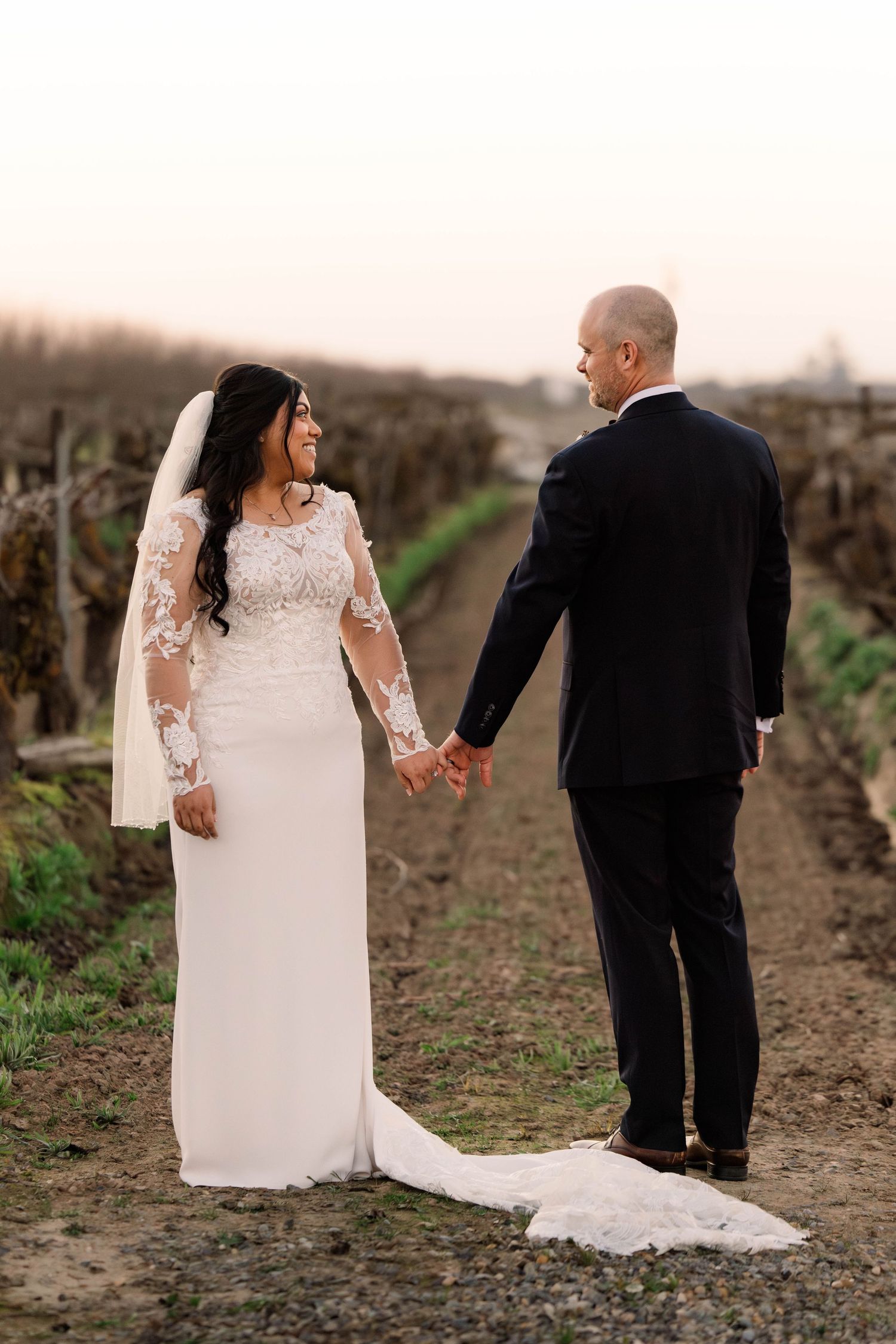 A couple holds hands while walking through vineyard rows at sunset.